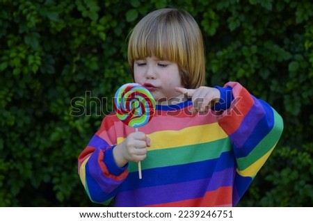 Funny kid with caramel on a stick. Cute positive boy 3 years old in a colorful t-shirt. Against the background of green grass. Space for text. Concept: kindergarten, candies, holidays, good mood