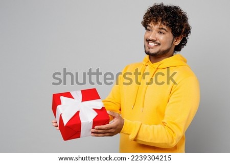 Side view young smiling happy Indian man 20s he wearing casual yellow hoody hold in hand giving red present box with gift ribbon bow isolated on plain grey background studio. People lifestyle portrait