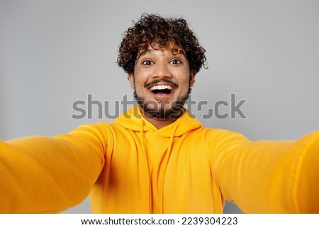Close up young cheerful happy fun Indian man 20s he wearing casual yellow hoody doing selfie shot pov on mobile cell phone isolated on plain grey background studio portrait. People lifestyle portrait