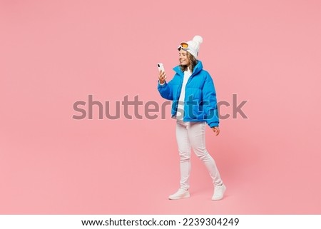 Snowboarder woman wear blue suit goggles mask hat ski padded jacket walk go use hold mobile cell phone isolated on plain pastel pink background. Winter extreme sport hobby weekend trip relax concept