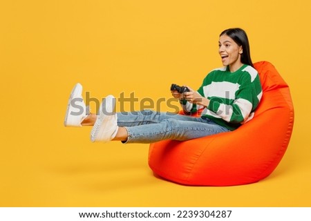 Full body young latin woman wear casual cozy green knitted sweater sit in bag chair hold in hand play pc game with joystick console isolated on plain yellow background studio People lifestyle concept