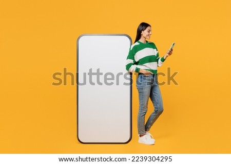 Full body fun young latin woman wear casual cozy green knitted sweater big huge blank screen mobile cell phone smartphone with area use smartphone isolated on plain yellow background studio portrait