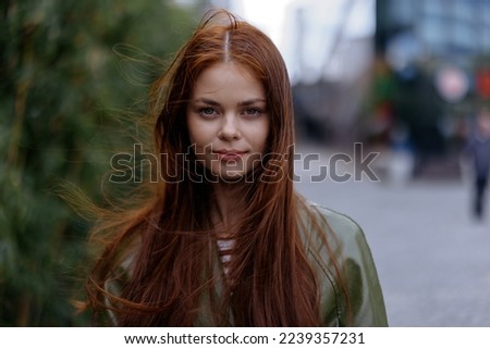 Portrait of a beautiful young girl in the city looking into the camera with red flying hair in a green raincoat in the city against a background of bamboo in spring, lifestyle in the city