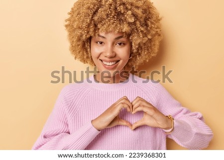 Positive curly haired woman smiles gently makes heart gesture says be my valentine expresses love to you wears casual pink knitted jumper isolated over beige background. Body language concept