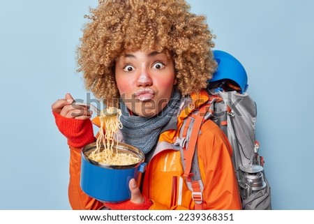Shocked curly woman camper eats noodles applies protective skin care cream carries rucksack with necessary equipment enjoys camping time isolated over blue background. Active journey concept