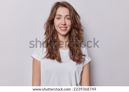 Portrait of beautiful blue eyed woman with dark wavy hair dressed in casual t shirt has natural beauty smiles toothily dressed in casual white t shirt isolated over greu background has healthy skin Royalty-Free Stock Photo #2239368749
