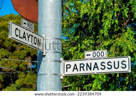 Road sign that says Clayton and Parnassus in San Francisco California with white and black paint on metal frame and pole. Green tree and foliage background in an urban area of the downtown city.