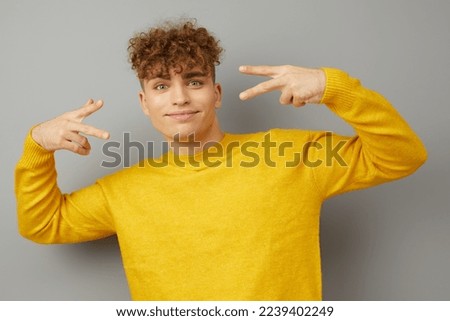 a pleasant joyful man stands on an isolated background in a bright yellow sweater and shows a peace sign with his fingers holding his hands near his head