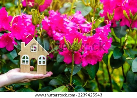 The girl holds the house symbol against the background of blossoming pink rhododendron
