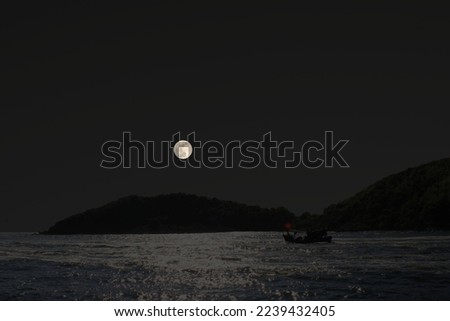 Photo of a fishing boat in front of an island in the sea at night at full moon.