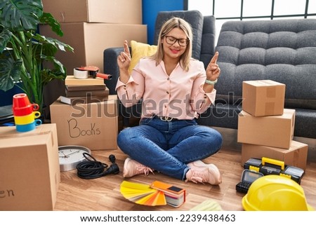Young hispanic woman moving to a new home sitting on the floor gesturing finger crossed smiling with hope and eyes closed. luck and superstitious concept. 