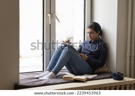Serious busy pretty Indian student woman studying at home, resting on soft windowsill with open book, copybook, using tablet for online Internet communication, reading on screen Royalty-Free Stock Photo #2239453963