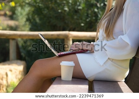 Detail of woman hands on laptop computer, next to her an eco-friendly paper cup of coffee or infusion. Woman is sitting on wooden bench. 