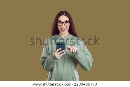 Joyfully excited female popular internet blogger enthusiastically checks comments under new post isolated on khaki background. Girl in casual hoodie happily smiling scrolling on mobile phone screen.