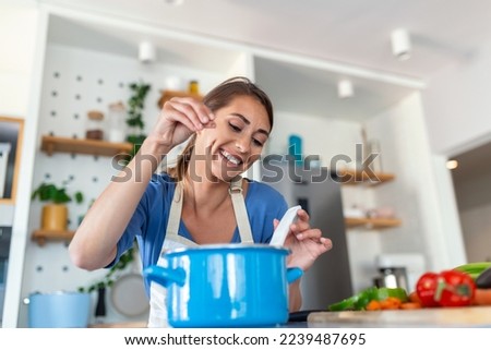 Happy Young Woman Cooking Tasting Dinner In A Pot Standing In Modern Kitchen At Home. Housewife Preparing Healthy Food Smiling . Household And Nutrition. Dieting Recipes Concept