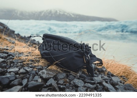 Close up camera bag on stones concept photo. Cameraman accessory outdoor. Front view photography with ice mountains on background. High quality picture for wallpaper, travel blog, magazine, article