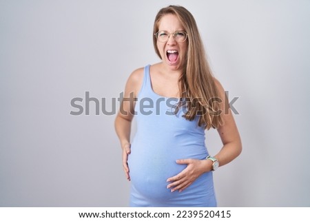 Young pregnant woman standing over white background angry and mad screaming frustrated and furious, shouting with anger. rage and aggressive concept. 