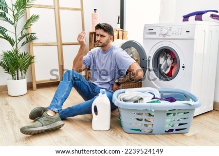 Young hispanic man putting dirty laundry into washing machine doing italian gesture with hand and fingers confident expression 