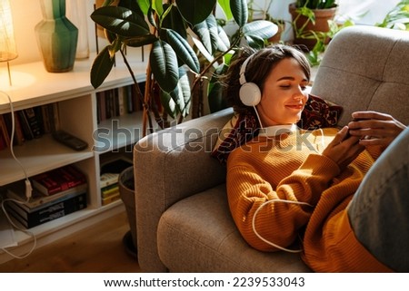 Brunette young woman listening music and using cellphone while resting on couch at home Royalty-Free Stock Photo #2239533043