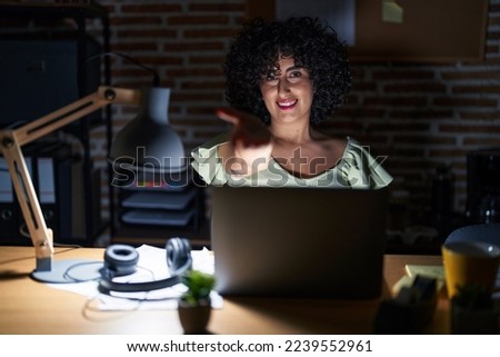 Young brunette woman with curly hair working at the office at night smiling cheerful offering palm hand giving assistance and acceptance. 