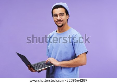 Young successful student or designer in blue t-shirt and beanie hat holding laptop while looking at camera against lavender background Royalty-Free Stock Photo #2239564289