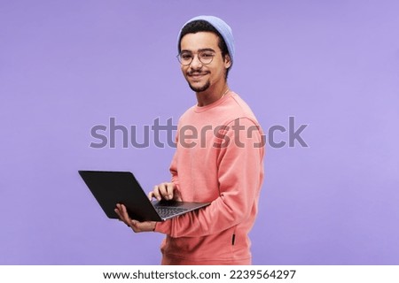 Young successful male freelancer or programmer in pink pullover, beanie hat and eyeglasses holding laptop and looking at camera Royalty-Free Stock Photo #2239564297