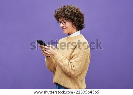 Side view of happy young brunette woman in eyeglasses and beige pullover using smartphone while standing on violet background