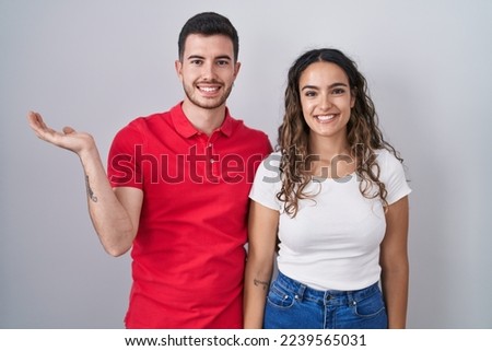 Young hispanic couple standing over isolated background smiling cheerful presenting and pointing with palm of hand looking at the camera. 