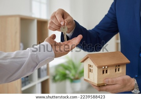 Real estate agent hands over key to customer after signing home purchase agreement. Close up of man's hand holding small wooden model house and handing keys to another man's hand. Real estate concept. Royalty-Free Stock Photo #2239588423