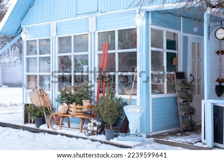 Interior home with big glass windows and wooden porch. Facade blue house with terrace in winter. Exterior Rustic country house with a gray roof. Snow Street with fir in pots, skis and sleds near house