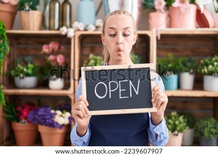 Young caucasian woman working at florist holding open sign making fish face with mouth and squinting eyes, crazy and comical. 