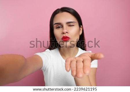Beautiful young woman taking selfie while blowing kiss on pink background, closeup
