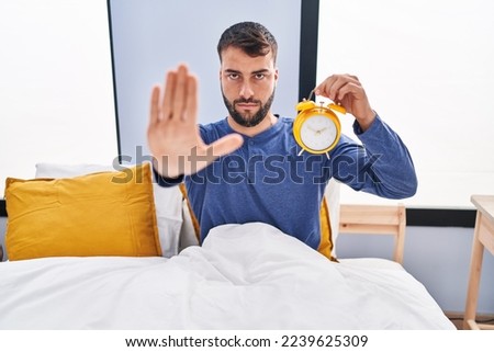 Handsome hispanic man in the bed holding alarm clock with open hand doing stop sign with serious and confident expression, defense gesture 