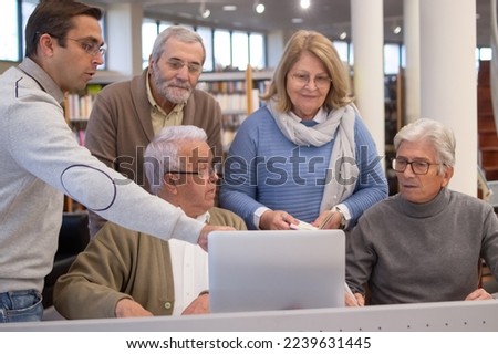 Portrait of group of aged people with teacher in library. Happy men and woman finding material for study in internet and young teacher explaining and helping them. Education for aged people concept Royalty-Free Stock Photo #2239631445
