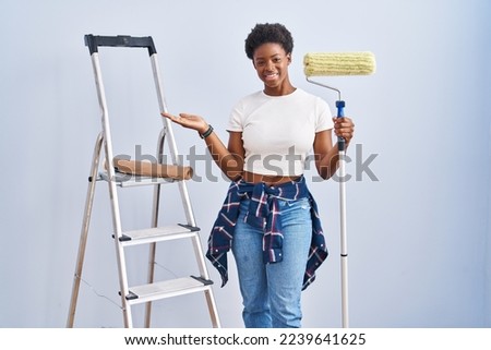 African american woman holding roller painter smiling cheerful presenting and pointing with palm of hand looking at the camera. 