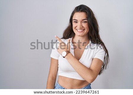 Young teenager girl standing over white background cheerful with a smile on face pointing with hand and finger up to the side with happy and natural expression 