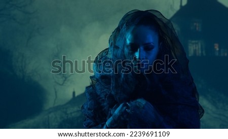 Beautiful witch making the witchcraft over the smoky background at night. Scary house on the hill. Halloween image.