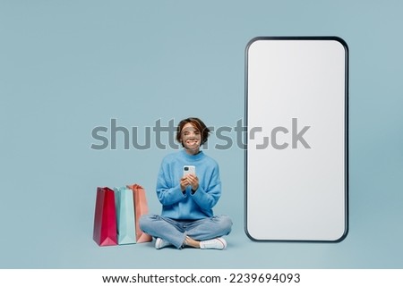 Full body young woman wear knitted sweater near package bags after shopping big huge blank screen mobile cell phone smartphone with workspace mockup area isolated on plain pastel light blue background