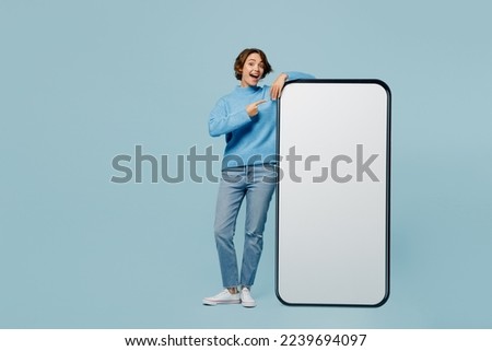 Full body amazed young woman wear knitted sweater point index finger big huge blank screen mobile cell phone smartphone with workspace mockup area isolated on plain pastel light blue cyan background