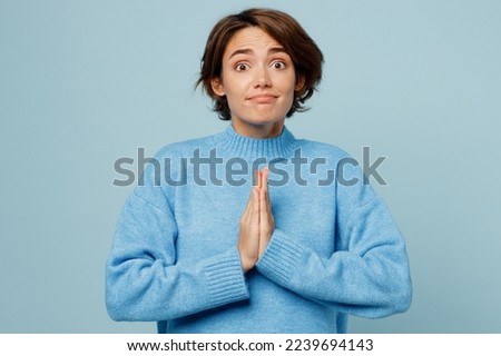 Young caucasian woman wear knitted sweater hold hands folded in prayer gesture, begging about something isolated on plain pastel light blue cyan background studio portrait. People lifestyle concept