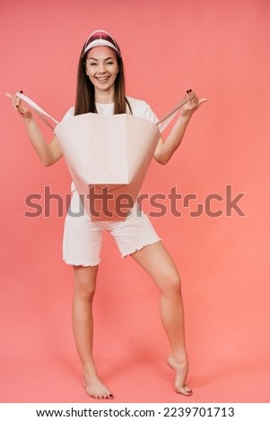 Vertical young Italian woman with long hair in a white T-shirt, colored transparent visor, holds open bag, looks at camera happily, after shopping On a pink background with an empty space for ad.