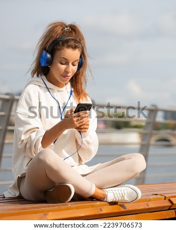 Outdoor vertical photo of a young woman in headphones with smartphone during a warm spring day. She smiles thoughtfully and looks away. Format photo 4x5.