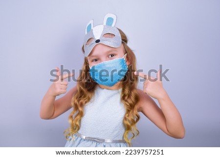 A child in a carnival masquerade mask of a rabbit hare and a medical mask decorated with rhinestones. Symbol of 2023. Mask fashion. Child is ready to celebrate safely