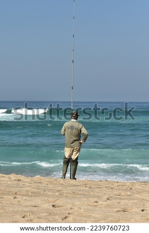 Wiry beach fisher in scruffy fishing outfit holding his rod at Praia da Foz do Arelho beach, Portugal, and observing the regular turquoise surf on a calm and sunny autumn day Royalty-Free Stock Photo #2239760723
