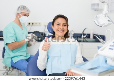 Happy cute latin woman sitting in dental chair after teeth cure giving thumb up Royalty-Free Stock Photo #2239761443