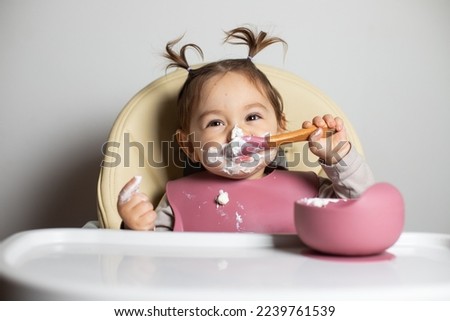 Small cute little toddler brunette caucasian girl with two tails tasting and enjoying by herself with a spoon the greek yogurt sitting in baby chair with messy face; self-feeding concept Royalty-Free Stock Photo #2239761539
