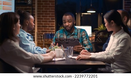 African american analytics specialist showing data visualization on laptop screen in business meeting. Teamwork, diverse coworkers discussing company report presentation at night time