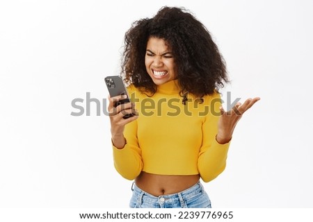 Shocked, angry african american girl, reacts on smth frustrating on mobile phone, complaining on smartphone text, standing over white background Royalty-Free Stock Photo #2239776965