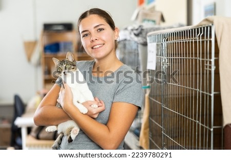 Happy caring young girl holding curious white and gray cat in arms while visiting shelter for abandoned animals. Pet adoption concept Royalty-Free Stock Photo #2239780291