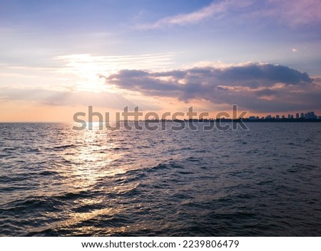 Autumn sunset view across stretch of water with waves beating boulders on the bank under blue sky, sun beaming between pink clouds creating light path on water